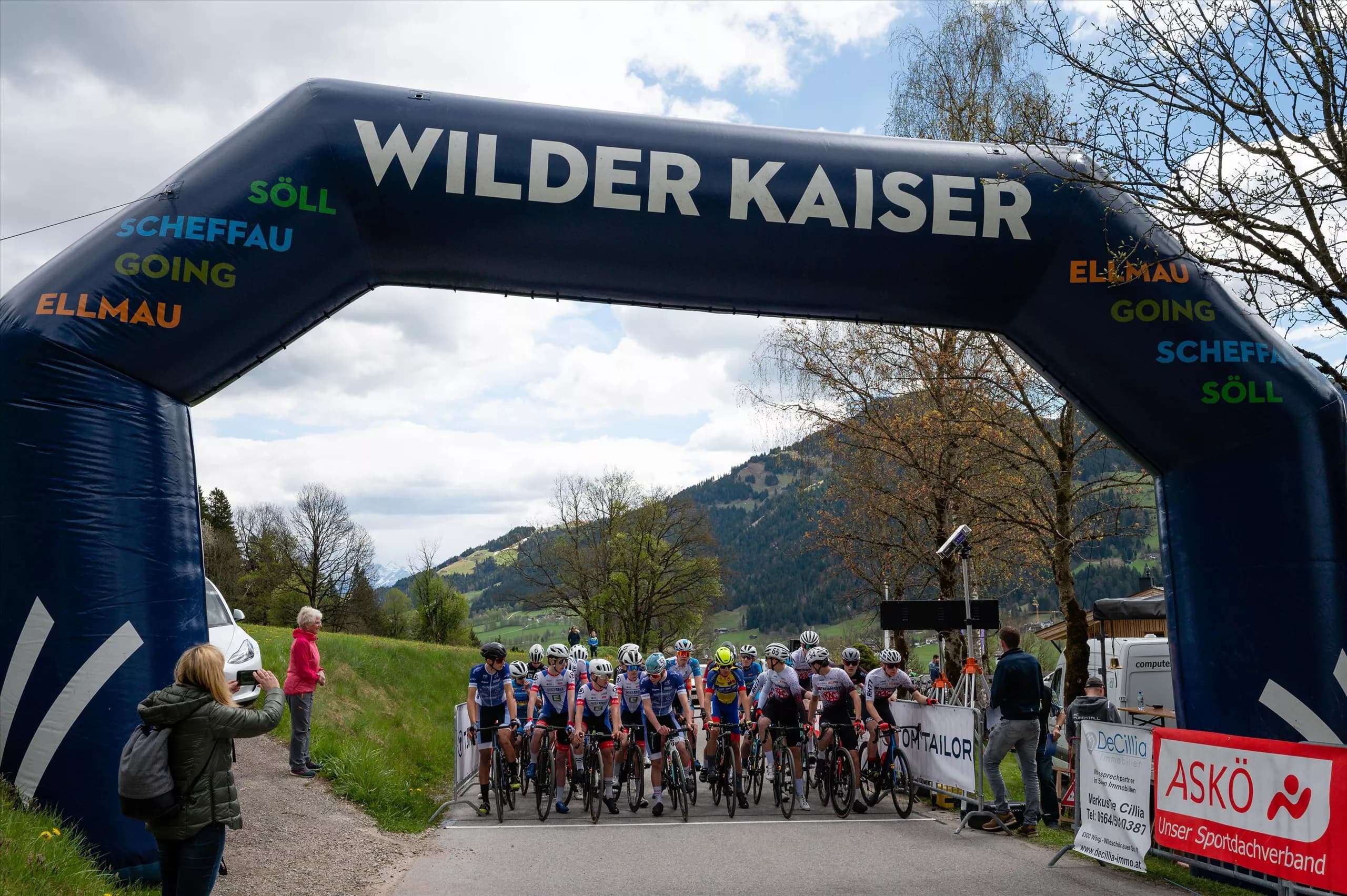 A bicycle race begins under a large arch with the inscription "Wilder Kaiser". In the background are mountain landscapes and the participants ready to start.