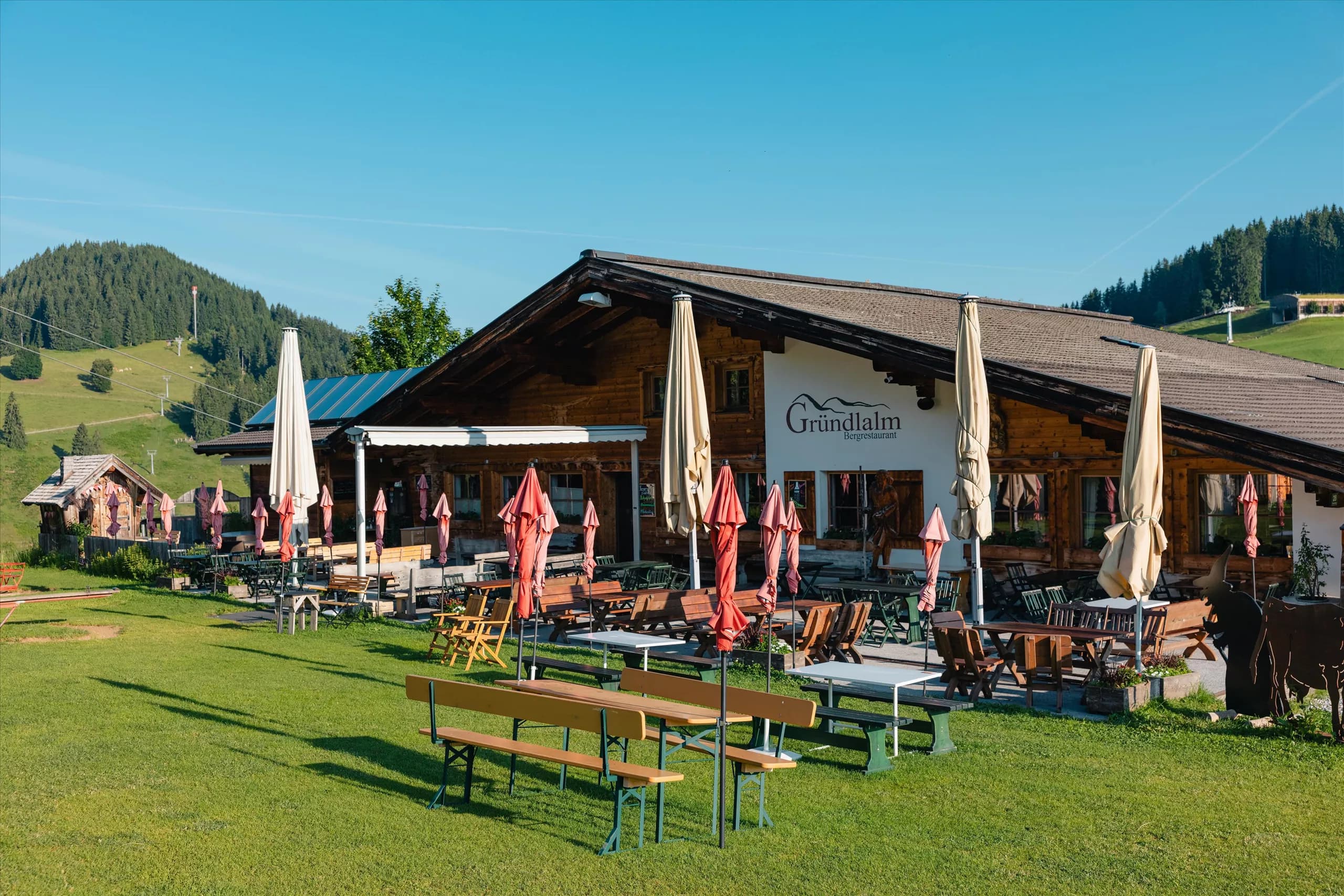 A cozy restaurant with an outdoor terrace, surrounded by meadows and mountains. Sunshades provide shade for the guests.