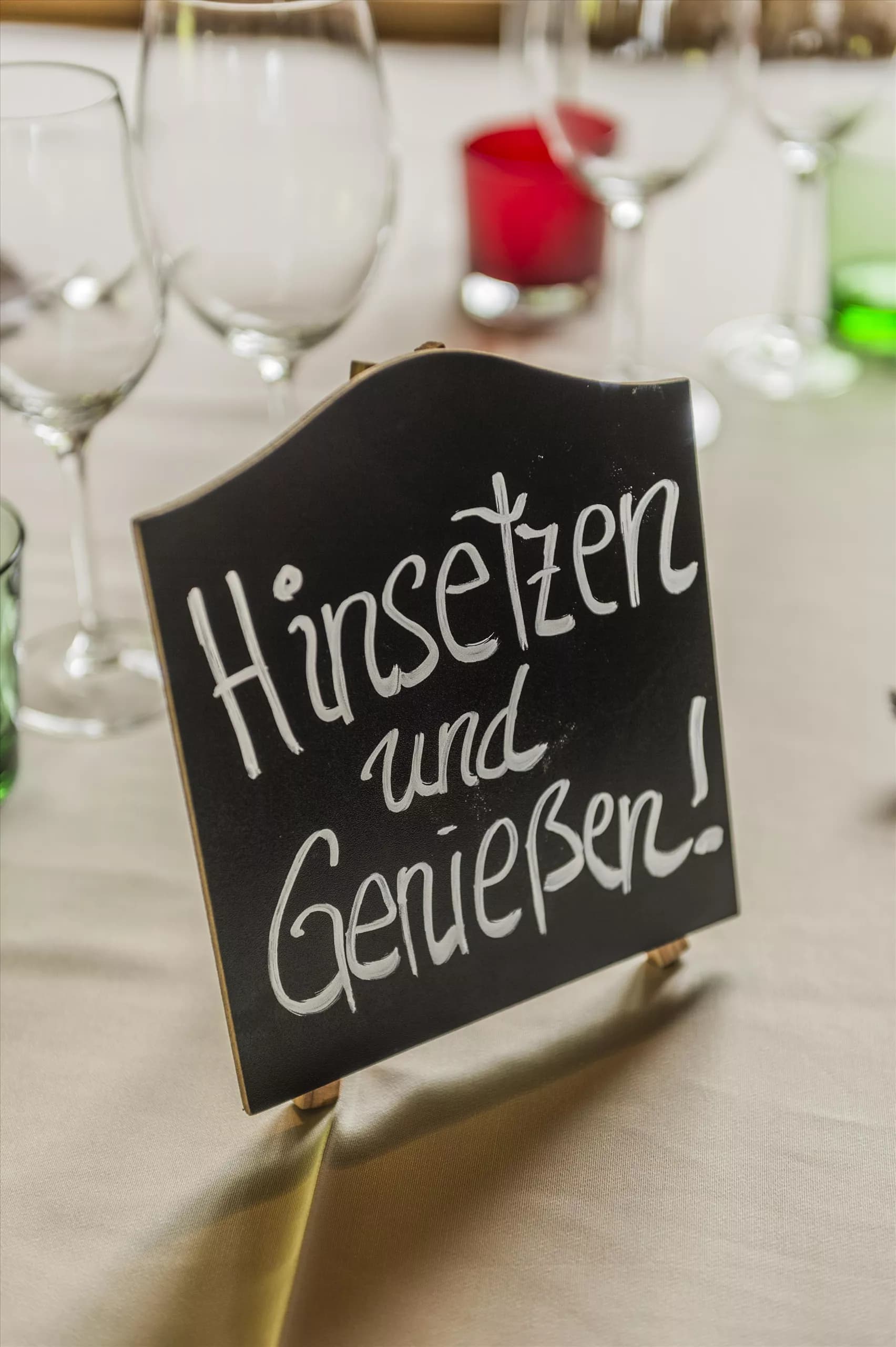 Ein stilvolles Tischsetting mit Gläsern und bunten Kerzen. Ein Schild lädt mit „Hinsetzen und Genießen!“ ein.