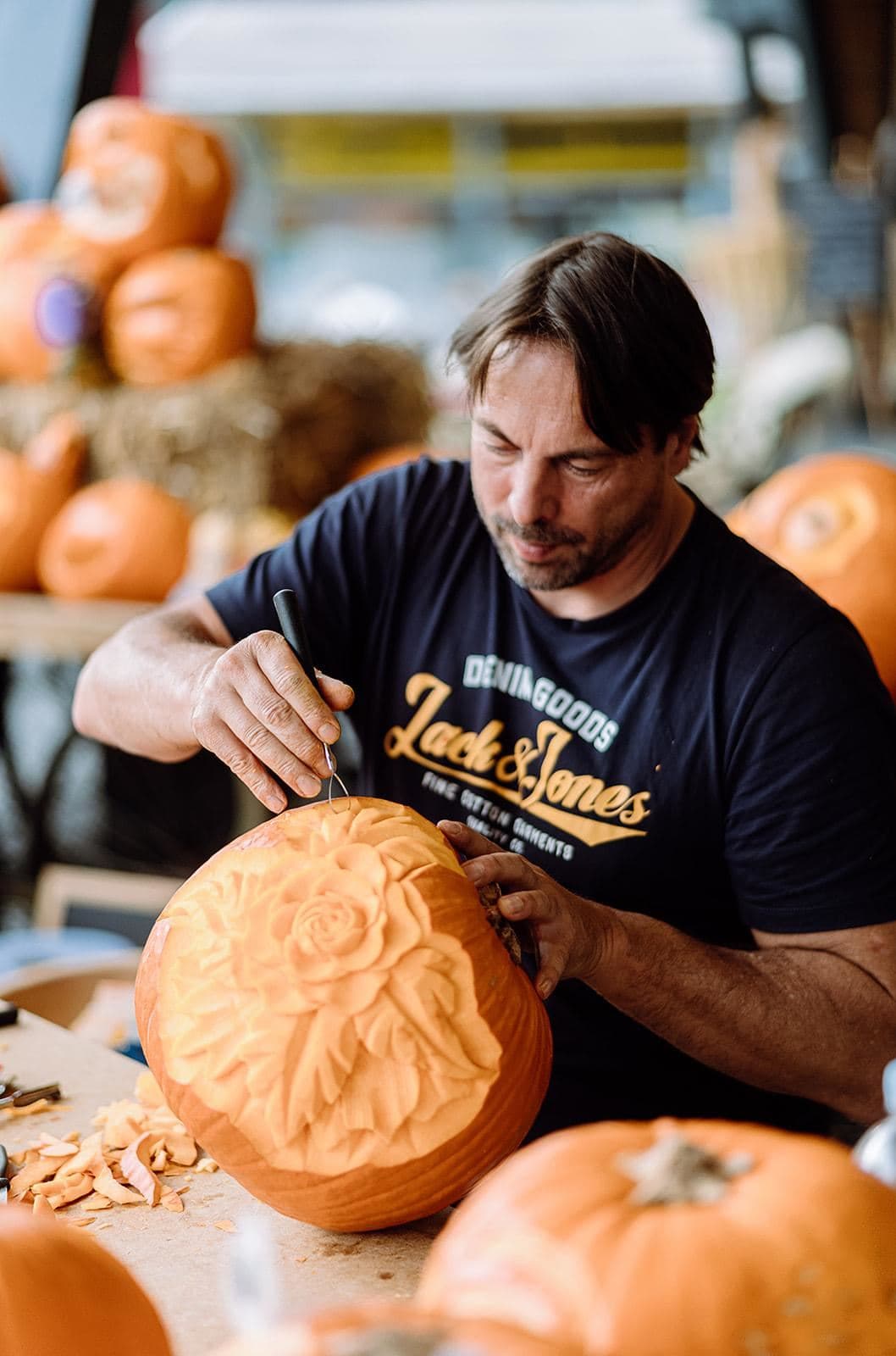 A man is carving a pumpkin with a tool. In the background, more pumpkins and straw bales are visible.