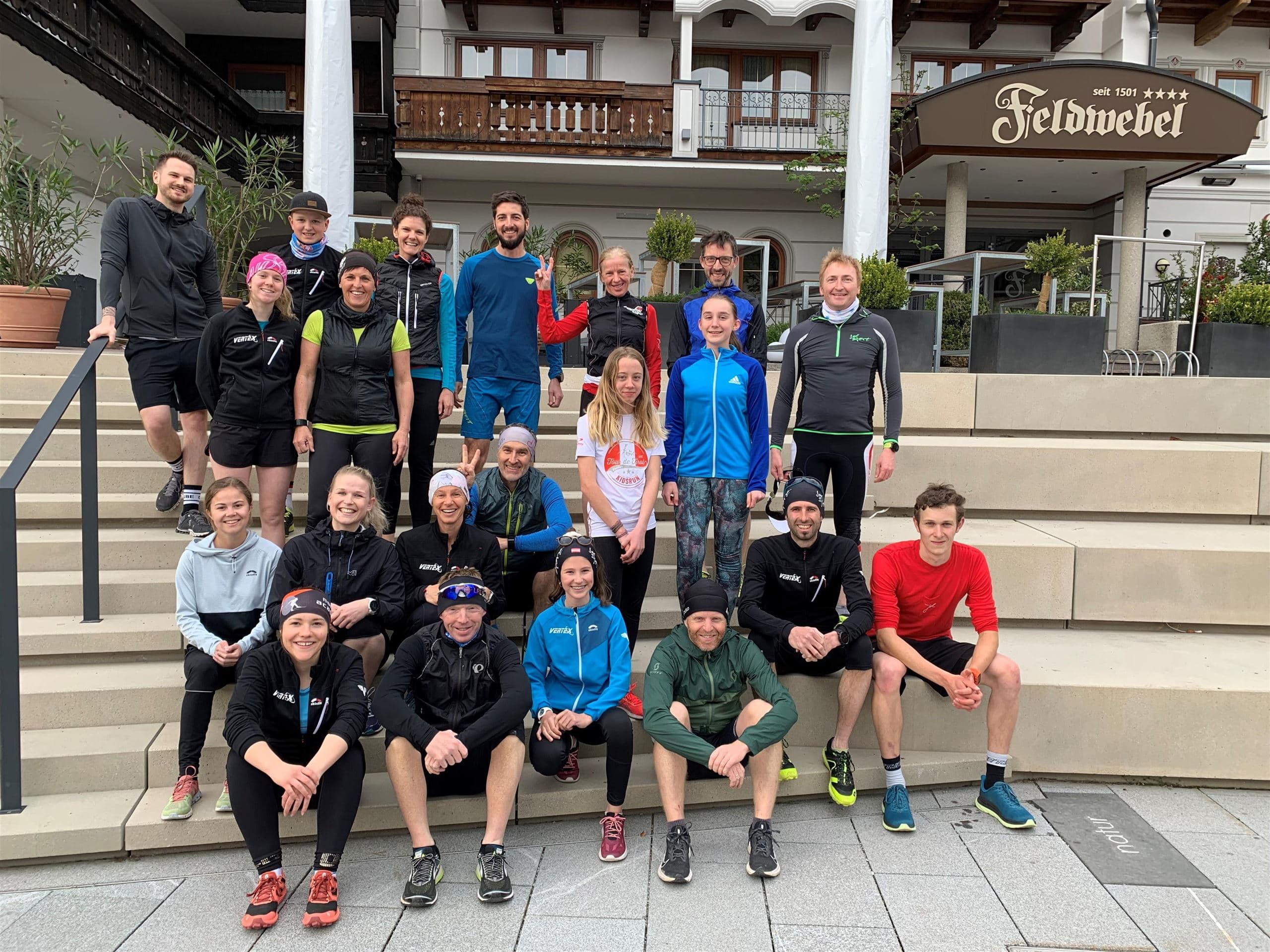 A group of people stands on the steps in front of a hotel. Everyone is dressed in sporty attire and is posing happily for the photo.