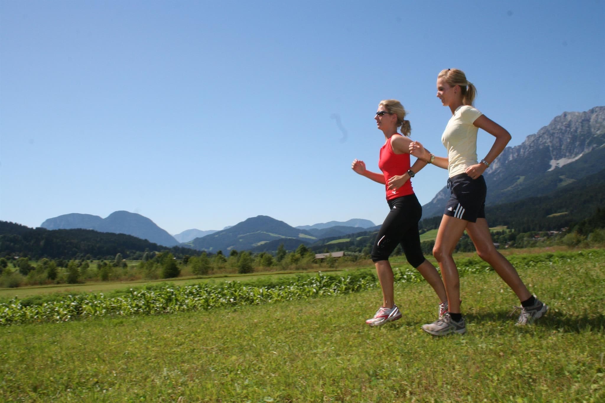 Two women are jogging on a green field with mountains in the background. The sky is clear and blue.