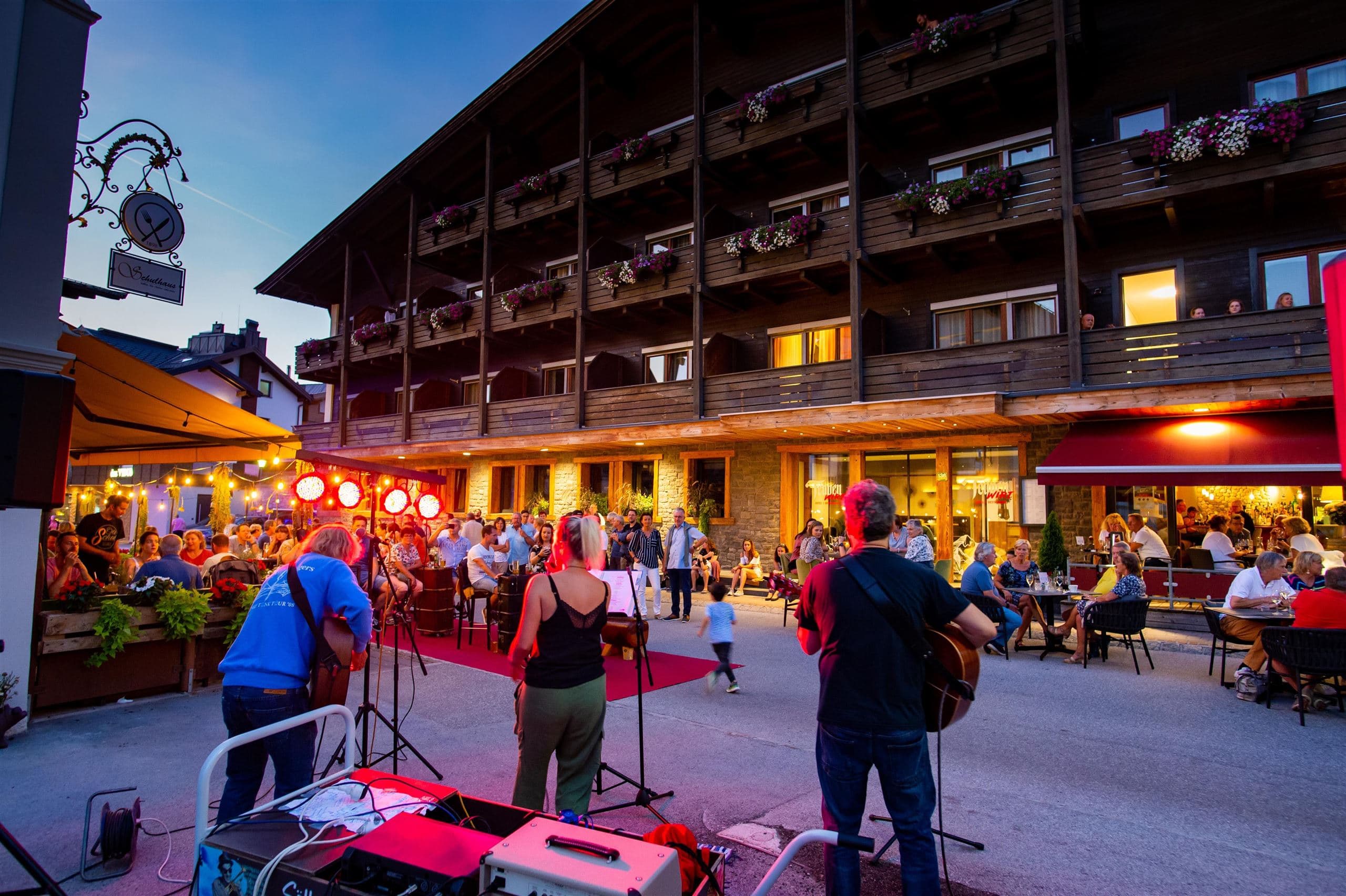 A busy street scene with musicians performing in front of a wooden building. Many people are sitting outside and enjoying the evening atmosphere.