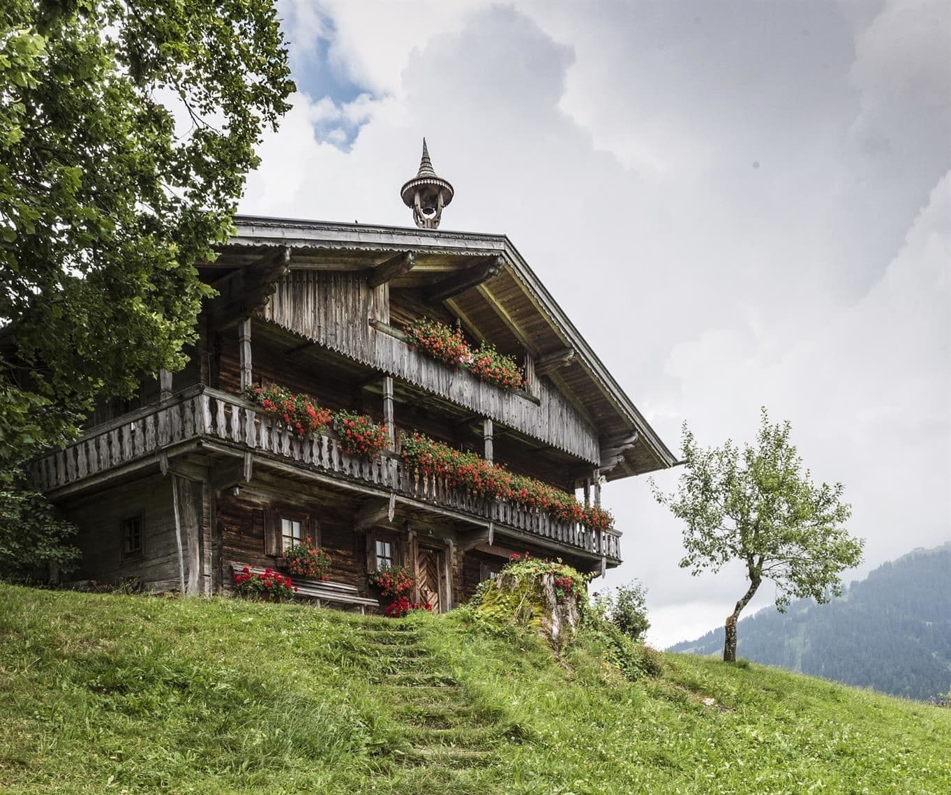 Ein traditionelles Holzhaus mit Balkon und bunten Blumen. Es steht in einer grünen Landschaft unter einem bewölkten Himmel.